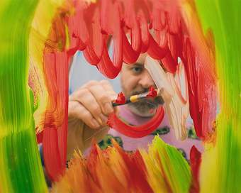 Bruce Asbestos looks through glass which he is painting on with red paint